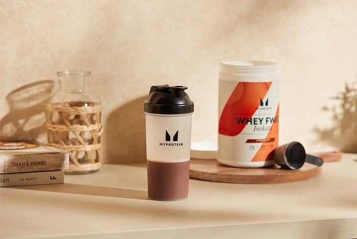 whey forward isolate chocolate flavour inside a shaker on a kitchen counter with a tub of protein powder behind