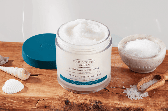 White container of the Cleansing Purifying Scrub with Sea Salt placed next to sea shells and salt on a wooden table.