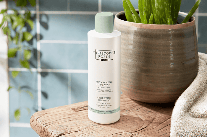 White bottle of the Hydrating Shampoo with Aloe Vera in a bathroom next to a plant on a wooden table.