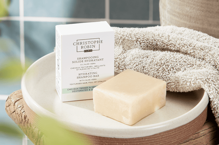 White box of Hydrating Shampoo Bar with Aloe Vera in a bathroom next to a beige towel on top of a wooden stool. 