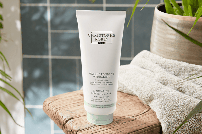 White bottle of the Hydrating Melting Mask with Aloe Vera next to a beige towel and green plant on top of a wooden stool.