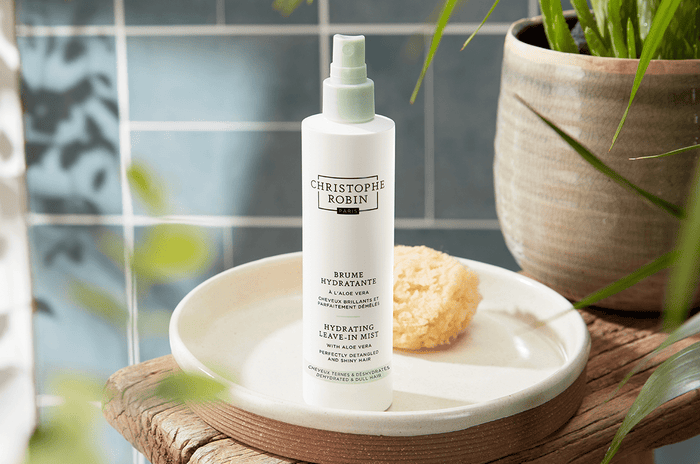 White bottle of the Hydrating Leave-in Mist with Aloe Vera next to a sponge and green plant on top of a wooden stool.