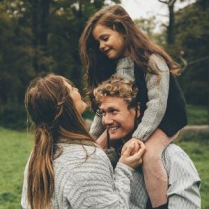 A daughter sat on her father's shoulders smiling at her mother outside.