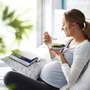 A pregnant woman in bed reading and eating a bowl of fruit and yogurt.