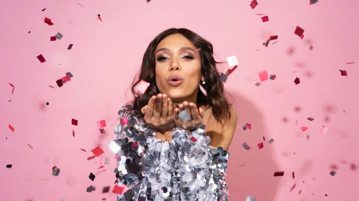 A woman in a sequined dress and subtle festive makeup blows a kiss while surrounded by confetti.