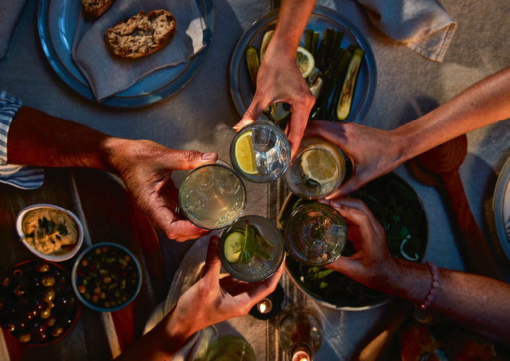 Bird's eye view of 5 people's arms reaching together to cheers drinks over a table of nibbles