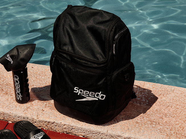 Shop swim accessories – black Speedo Teamstar rucksack on pool bench filled with swimming accessories.