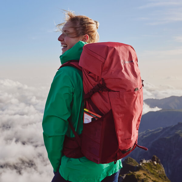 A Female stood at the top of the mountain overlooking the views whilst showcasing a red Begrhaus rucksack, on the left of the image in white text it says "why w do what we do (and why we love it)