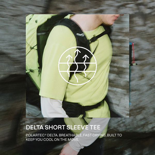   A person hiking outdoors, wearing a light green short-sleeve T-shirt over a long-sleeve beige base layer. They carry a large light green hiking backpack with black straps and attachments, including trekking poles secured to the side. The person also wears a black cap and dark pants, moving along a rocky surface.- WITH TEXT "POLARTEC® FABRICS DELIVER ADVANCED THERMAL REGULATION, BREATHABILITY AND DURABILITY. BUILT TO KEEP YOU MOVING IN ANY CONDITION." 