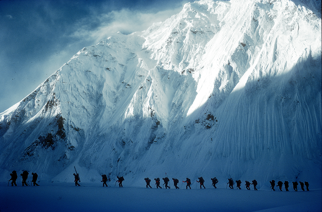 an image of a big snowy mountain with the silhoutte of a line of people walking across the bottom of the mountain