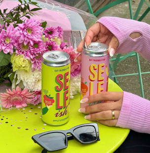 A hand in the middle of opening a can of the Peach & Orange Sparkling Prebiotic Drink next to a can of Apple & Raspberry Sparkling Prebiotic Drink, on a green table with flowers and sunglasses. Visit us on Instagram at selfishdrinks.