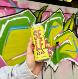 An arm holding a can of the Apple & Raspberry Sparkling Prebiotic Drink, in front of a graffitied wall. Visit us on Instagram at selfishdrinks.