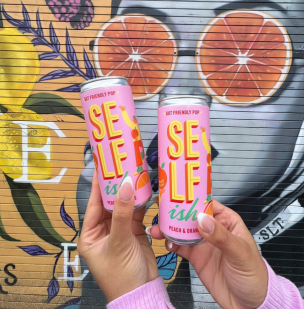 Two hands holding two cans of the Peach & Orange Sparkling Prebiotic Drink, in front of a graffitied wall. Visit us on Instagram at selfishdrinks.