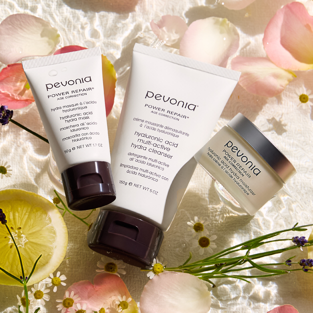 Pevonia's new Hyaluronic Acid products - a cleanser, mask and moisturizer lay on a bed of water surrounded by the product ingredients, including lemon slices, Lavendar and rose petals 