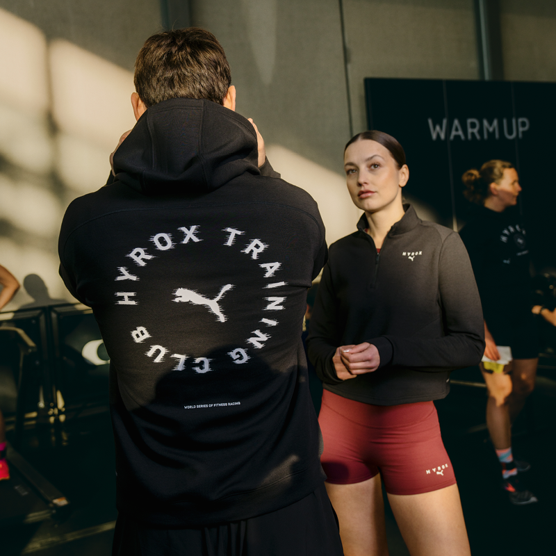Man with back towards camera, showing off his HYROX hoodie, with woman infront of him facing the camera, showing her merch.