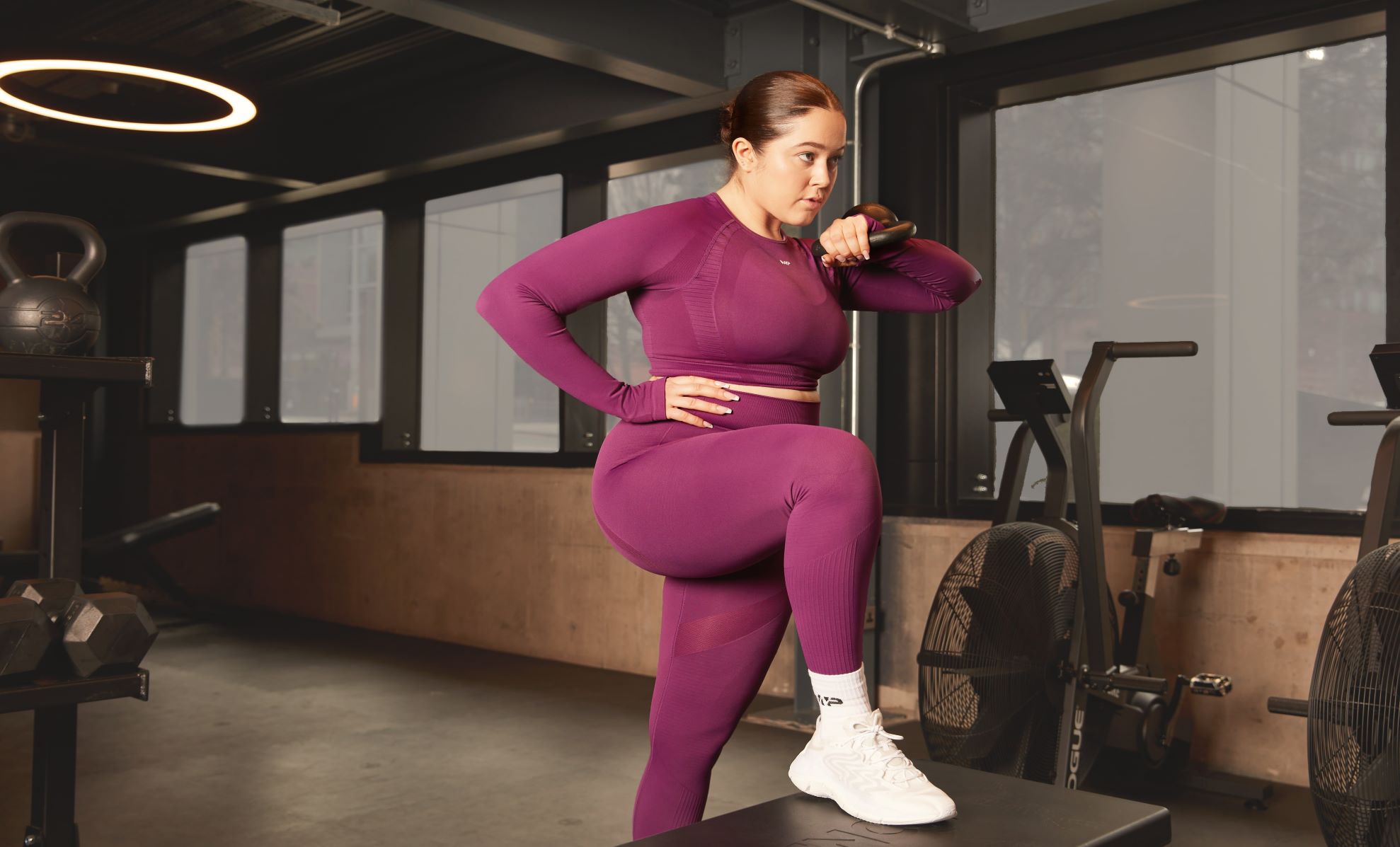 A woman in pink MP gym clothing working out in a gym with a black kettlebell.