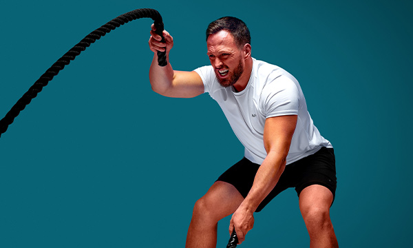 A man in a white t-shirt with the MP logo and black gym shorts working out with battle ropes on a light blue background.