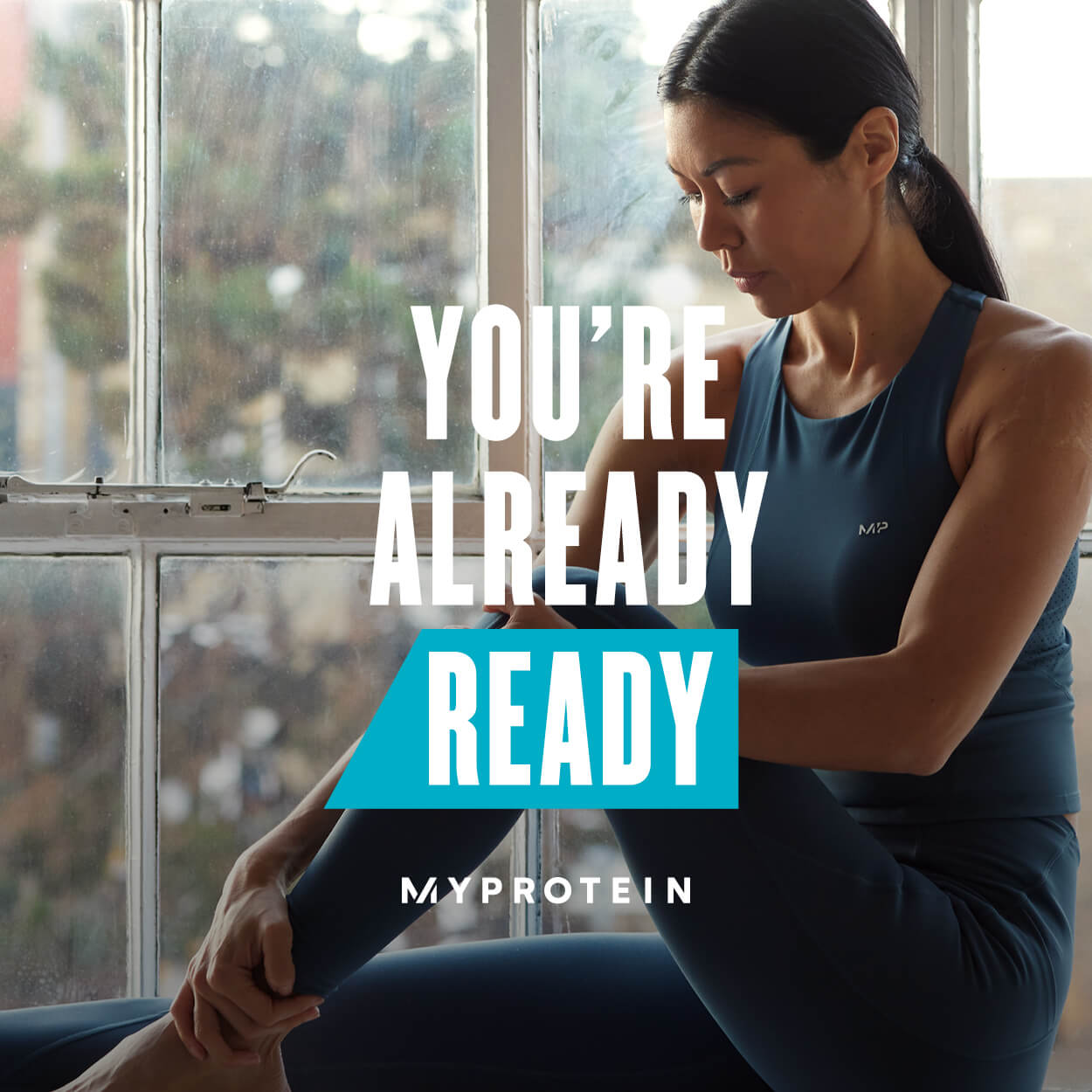 A woman wearing MP gym clothing sat on a window ledge stretching with the text 'You're Already Ready - MYPROTEIN' overlaying the image.